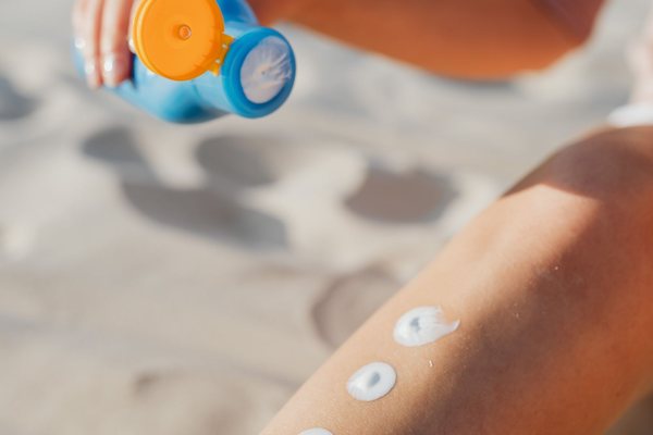 A woman applying sunscreen lotion from a bottle onto her leg while sitting on the sand at the beach.