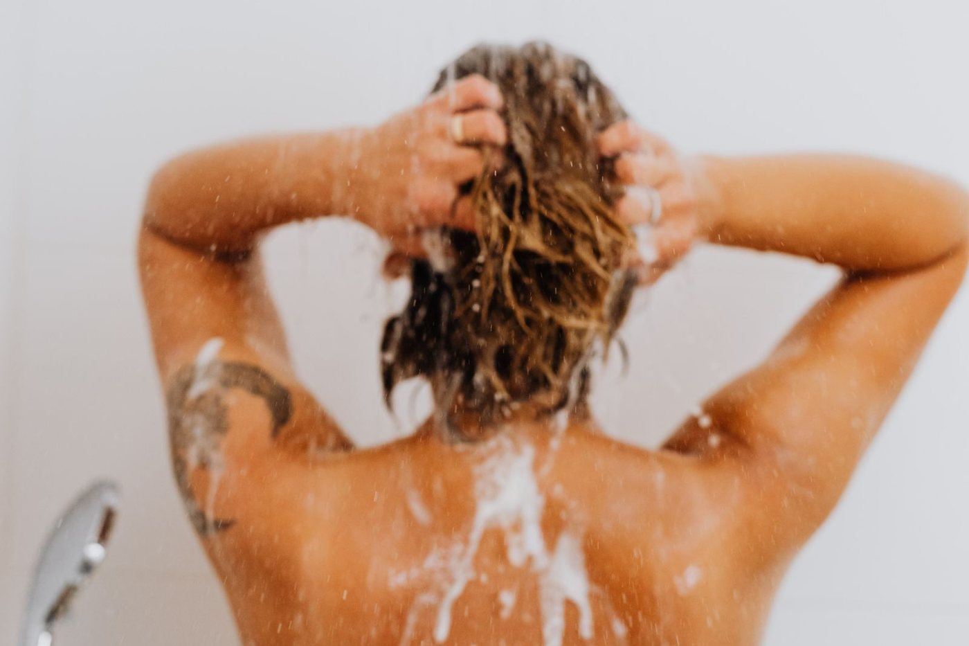 Back view of a woman lathering shampoo through her hair while washing in the shower.